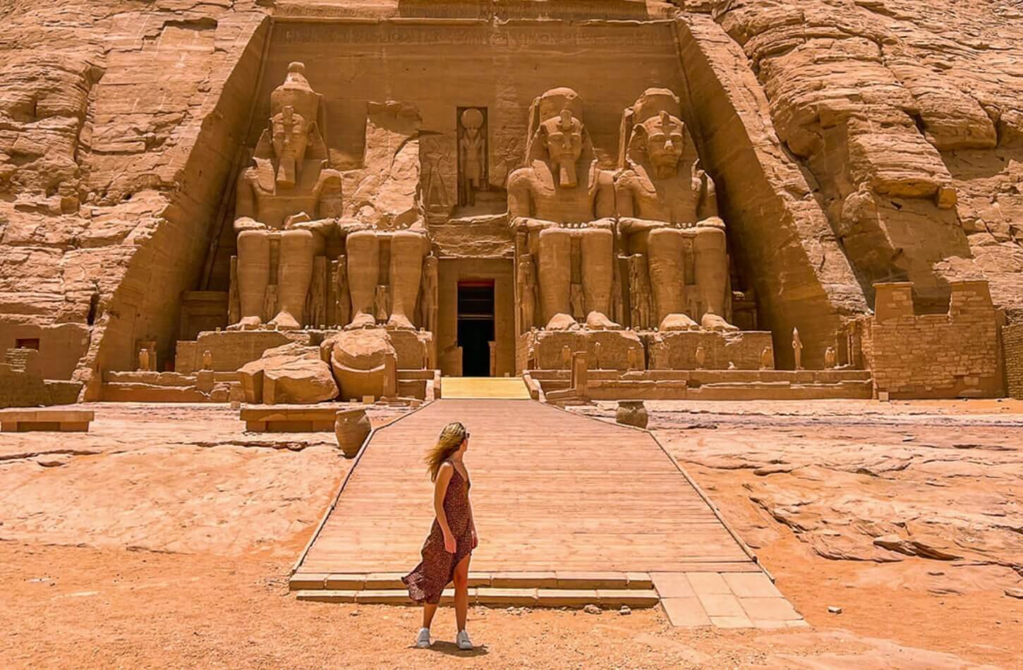 Is It Safe to Travel to Egypt? a woman walks towards the massive statues of abu simbel dressed in a light airy dress and bathed in golden sunlight