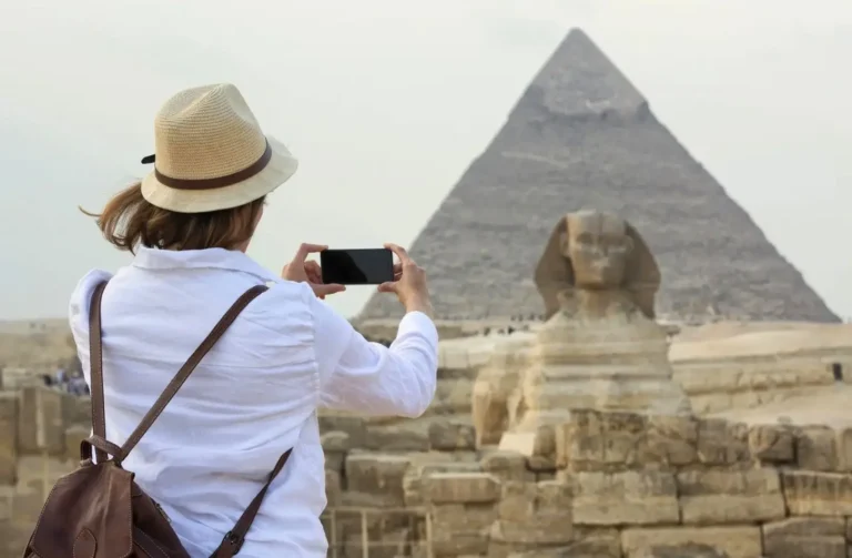 Egypt itinerary 7 days featuring a female traveler in a white shirt and sun hat taking a photo of the Great Sphinx and the Pyramid of Khafre with her smartphone at the Giza Plateau.