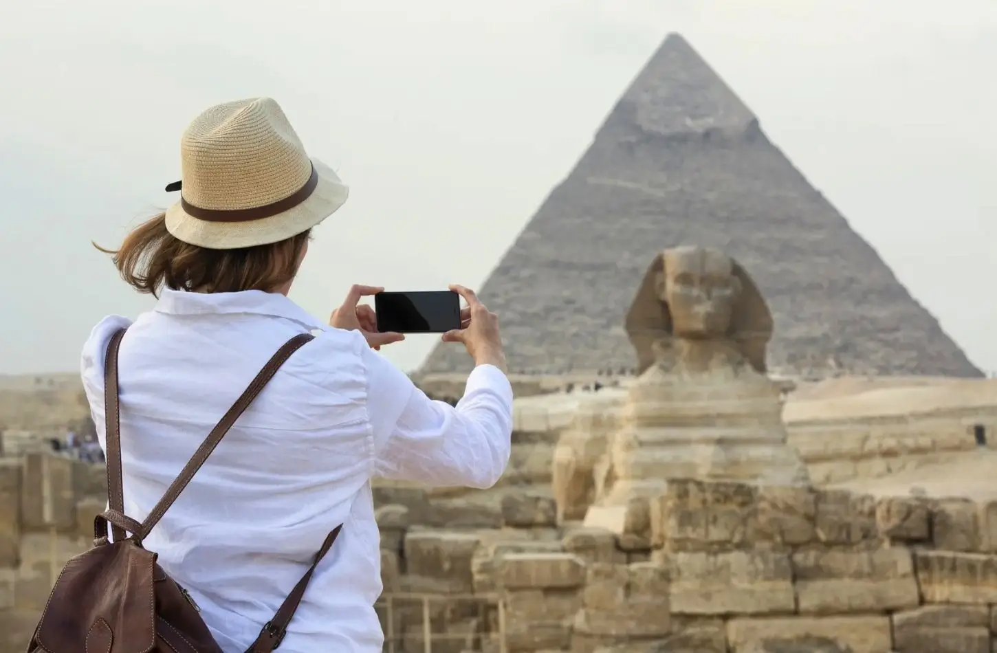 Egypt itinerary 7 days featuring a female traveler in a white shirt and sun hat taking a photo of the Great Sphinx and the Pyramid of Khafre with her smartphone at the Giza Plateau.