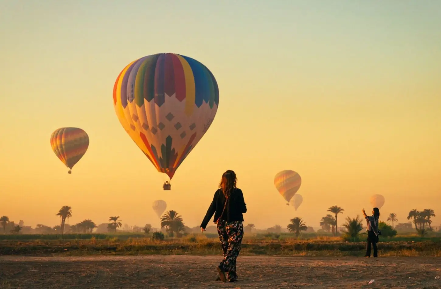 A woman in patterned pants walks through a dusty field during her solo female travel in Egypt, watching colorful hot air balloons float over a hazy landscape with palm trees at sunrise.