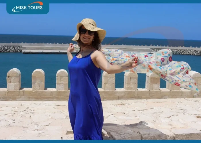 A-smiling-woman-in-a-blue-dress-and-sun-hat-holding-a-flowing-scarf-by-the-sea.