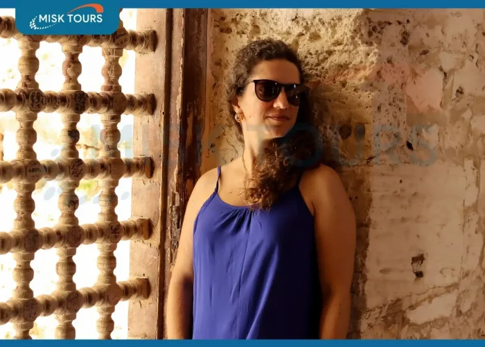a-woman-in-a-blue-dress-and-sunglasses-leaning-against-a-stone-wall-by-a-wooden-mashrabiya-window.