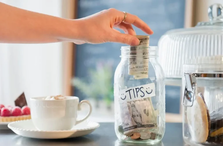 A hand placing cash into a glass jar labeled "TIPS" on a counter, representing the culture of Tipping in Egypt.