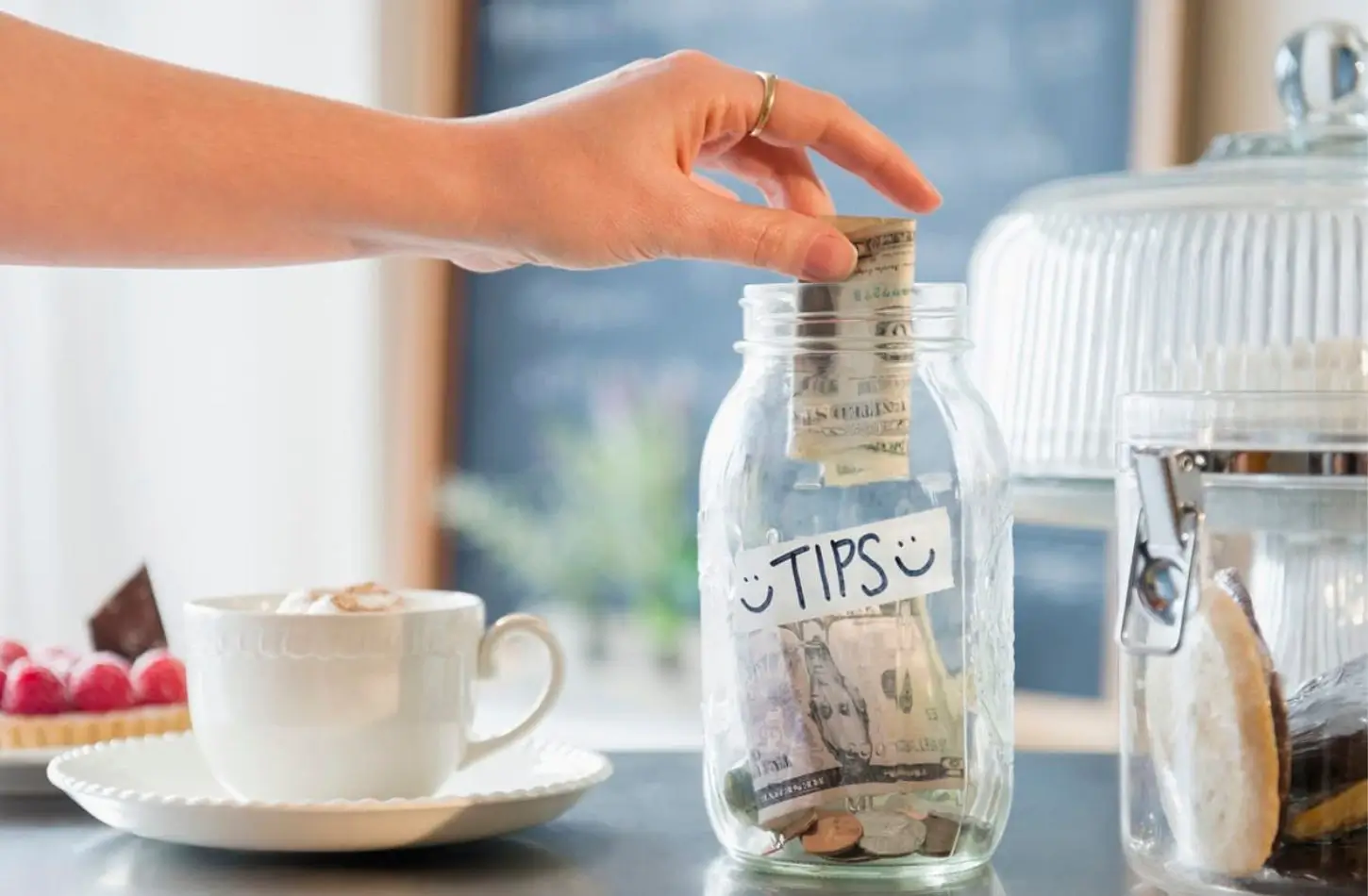 A hand placing cash into a glass jar labeled "TIPS" on a counter, representing the culture of Tipping in Egypt.
