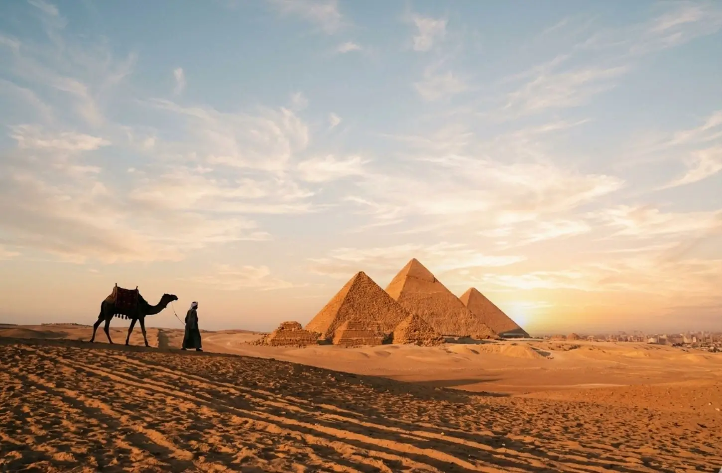 Egypt in December sightseeing view showing a camel handler leading a camel across desert sand dunes at sunset, with the three Pyramids of Giza standing against a soft blue and orange sky.