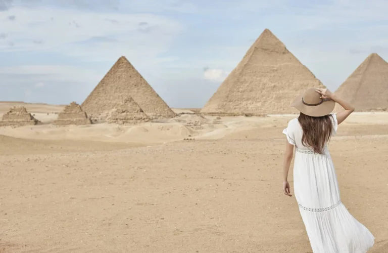 Female tourist in a white dress and sun hat enjoying clear spring weather at the Giza Pyramids while visiting Egypt in April.