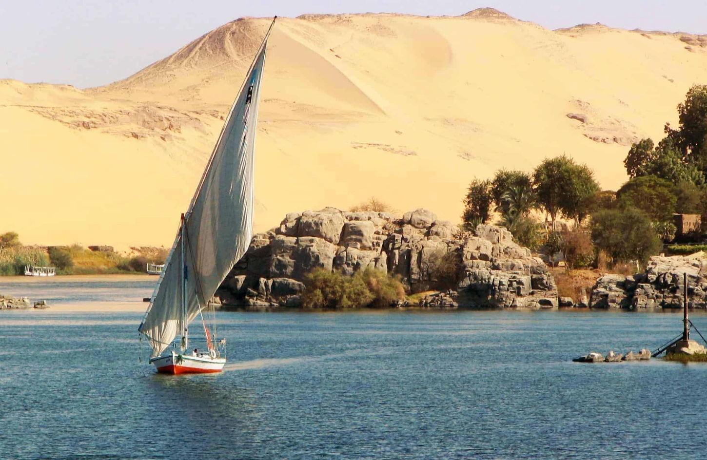 Traditional Egyptian felucca sailboat gliding along the Nile River past golden sand dunes while traveling in Egypt in April.